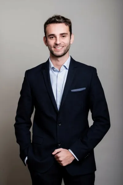 Homme souriant en costume bleu marine avec pochette, posant sur fond gris.