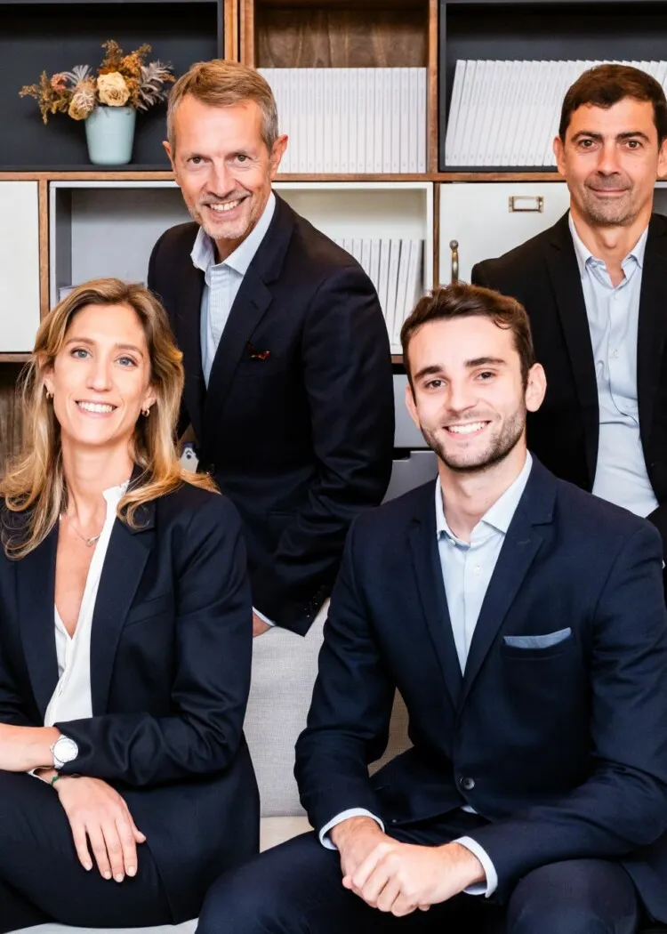 Quatre professionnels en costumes sombres souriant dans un bureau moderne.